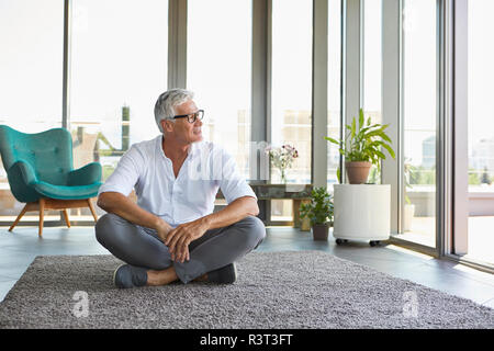 Homme mûr assis sur un tapis à la maison à la fenêtre de Banque D'Images