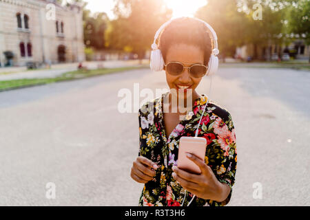 Heureux à la mode jeune femme avec un casque et smartphone outdoors at sunset Banque D'Images