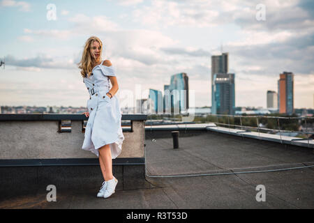 Jeune femme blonde debout sur la terrasse du toit wearing white dress Banque D'Images