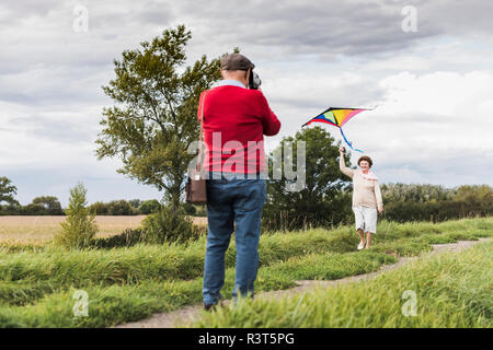 Man filming épouse flying kite in rural landscape Banque D'Images