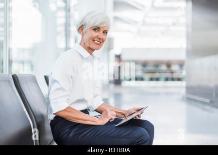 Smiling senior businesswoman sitting in waiting area en utilisant tablet Banque D'Images