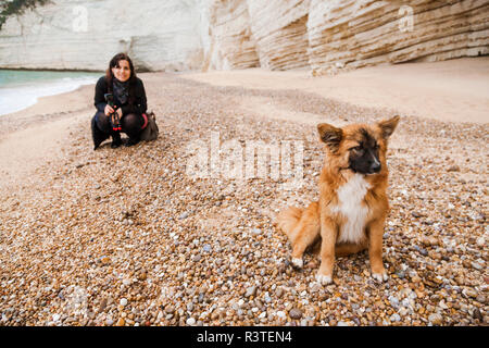 L'Italie, Vieste, stray dog sitting on Vignanotica plage tandis que smiling woman crouching dans l'arrière-plan Banque D'Images