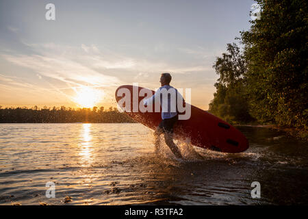 Homme avec paddleboard dans le lac à pied par le coucher du soleil Banque D'Images