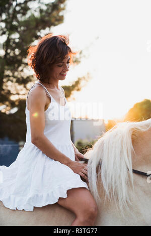 Smiling woman in white dress riding bareback sur le cheval au coucher du soleil Banque D'Images