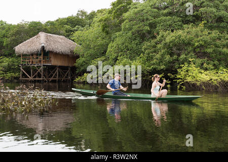 Les touristes en canot la rivière amazonienne près d'un toit traditionnel en chaume construction de huttes sur pilotis. (MR) Banque D'Images