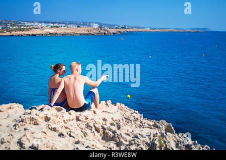 Couple assis ensemble sur un rocher près de l'eau transparente. L'homme est montrant quelque chose à une femme, en pointant. Concept d'amour, de soins, de voyager tog Banque D'Images