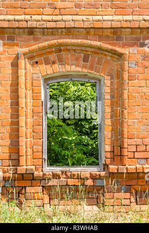 Fenêtre de l'ancien bâtiment détruit de brique rouge à travers lequel vous pouvez voir des plantes vertes et de l'herbe. Banque D'Images