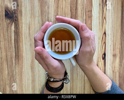 Un gros plan d'une woman's hands holding une tasse de thé chaud. L'arrière-plan est une table en bois. L'un des bracelets est en cuir et l'autre est silv Banque D'Images