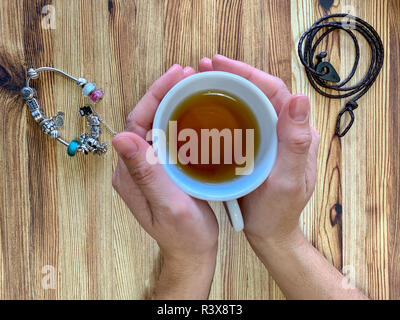 Un gros plan d'une woman's hands holding une tasse de thé chaud. L'arrière-plan est une table en bois. L'un des bracelets est en cuir et l'autre est d'argent Banque D'Images