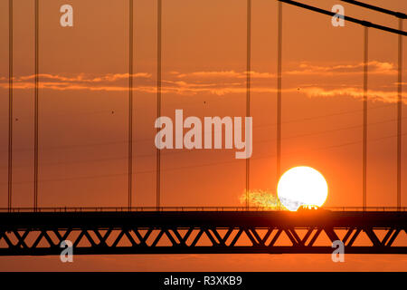 Une voiture roulant dans le soleil sur un pont suspendu et apparemment prendre feu lors d'un coucher de soleil rouge. Emmerich, Allemagne. Banque D'Images