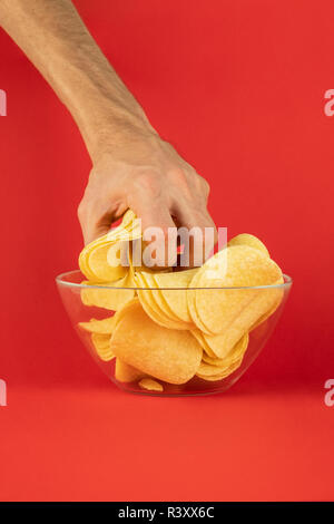 Accaparement des croustilles de pommes de terre à partir d'un bol en verre. Main mâle prend goulûment poignée de chips à l'arrière-plan rouge vif Banque D'Images