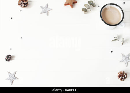 Composition de Noël. Branches d'eucalyptus, de stars, pommes de pin, des bâtons de cannelle et de confettis scintillants sur table en bois blanc, arrière-plan. Le café dans une tasse de l'émail. Petit-déjeuner d'hiver. Mise à plat, vue du dessus. Banque D'Images