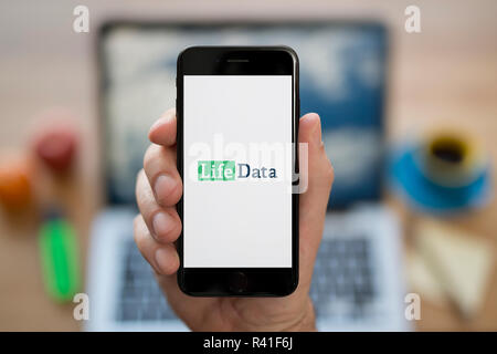 Un homme se penche sur son iPhone qui affiche les données de la vie, tandis que le logo était assis à son bureau de l'ordinateur (usage éditorial uniquement). Banque D'Images