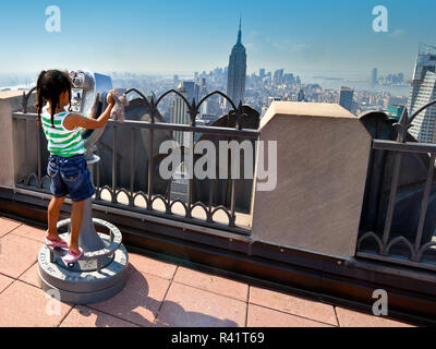 Une jeune fille avec un ours en peluche se tient sur la pointe des pieds pour regarder à travers les jumelles et la vue sur les toits de la ville de New York, à partir de la plate-forme d'observation au sommet Rockefel Banque D'Images