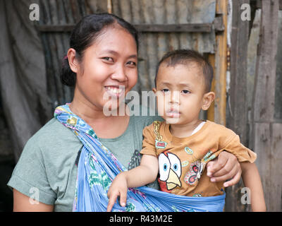 Portrait of smiling horizontale, fier, heureux jeune mère indonésienne holding Young boy wearing yellow t-shirt en face de la maison bois et de tôle. Banque D'Images