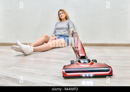 Fatigué femme de ménage est assis sur le plancher et nettoyer la poussière avec l'aspirateur vertical rouge moderne avec des lumières LED. Banque D'Images