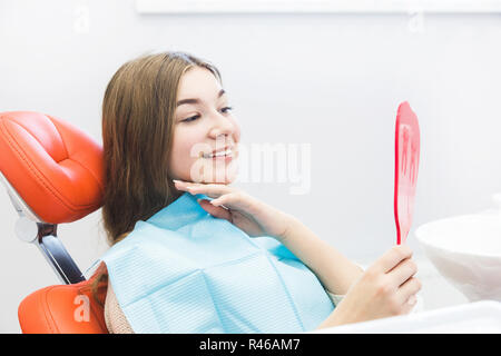 Clinique dentaire. La réception, l'examen du patient. Soins des dents. Jeune fille souriante, à la recherche dans le miroir après un bilan dentaire à son dentiste Banque D'Images