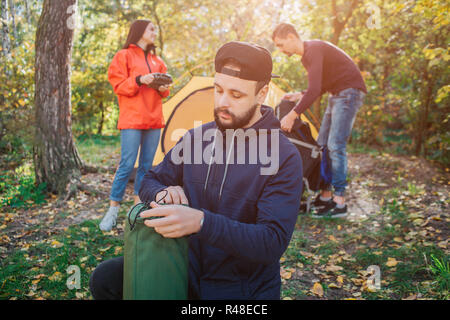 Jeune homme barbu est titulaire d'un sac de couchage et regarde. Il est sérieux. Femme détient la radio dans les mains. Un autre gars travaille avec tente. Banque D'Images