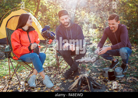 Les amis assis à feu et passer du temps ensemble. Jeune homme de droite contient grill avec les saucisses sur le feu. Ils sont la cuisine et le tabagisme. Un autre gars regarde. Jeune femme sourire. Banque D'Images