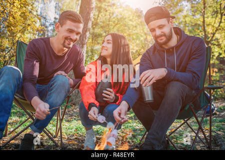 Heureux amis assis ensemble et murshmellow la cuisson sur le feu. Elle regarde guy à gauche et rire. Les jeunes hommes, l'air au feu. Certains d'entre eux ont des thermos dans les mains. Banque D'Images