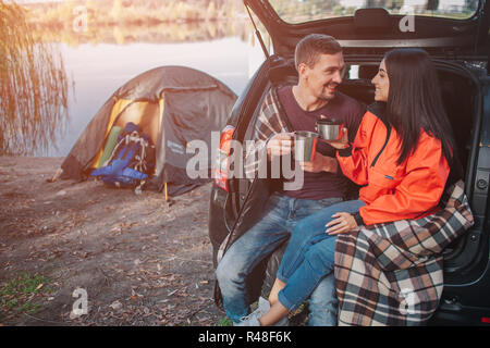 Image positive de cheerful couple sitting in trunk et regarder les uns les autres. Ils sourient et maintenez-tasses. Les jambes de la jeune femme sont couverts d'une couverture. Ils sont au bord du lac. Banque D'Images