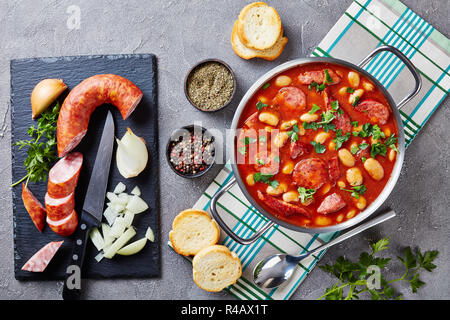 Ragoût de haricots avec des saucisses, des herbes et épices dans la sauce tomate dans une casserole de métal sur une table en béton avec des ingrédients, fasolka po bretonsku, polonais c Banque D'Images