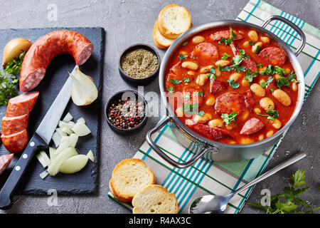 Ragoût de haricots avec des saucisses, des herbes et épices dans la sauce tomate dans une casserole de métal sur une table en béton avec des ingrédients, fasolka po bretonsku, polonais cui Banque D'Images