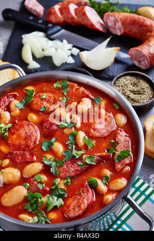 Ragoût de haricots copieux avec des saucisses, des herbes et épices dans la sauce tomate dans une casserole de métal sur une table en béton avec des ingrédients, fasolka po bretonsku, com Banque D'Images