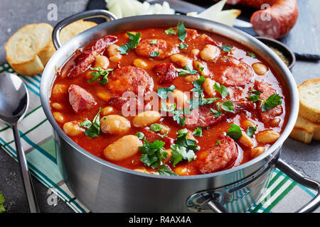 Ragoût de haricots copieux avec des saucisses, des herbes et épices dans la sauce tomate dans une casserole de métal sur une table en béton avec des ingrédients, fasolka po bretonsku, pol Banque D'Images