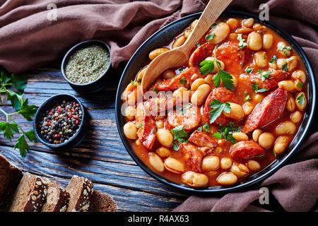 Ragoût de haricots copieux avec des saucisses, des herbes et épices dans la sauce tomate dans un bol noir avec une cuillère, sur une table en bois rustique avec un tissu marron, close-up Banque D'Images