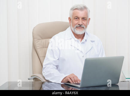 Le meilleur médecin senior working on laptop dans son bureau. Beau vieil homme vêtu en uniforme blanc, assis à table dans un fauteuil confortable et à la recherche à l'appareil photo. La médecine et les soins de concept. Banque D'Images