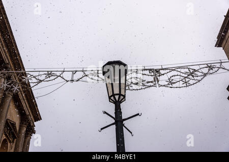 Lampe décorative d'un réverbère en face de décorations de fête entre 2 bâtiments avec un ciel gris blanc de jour avec (faux) la neige qui tombe Banque D'Images