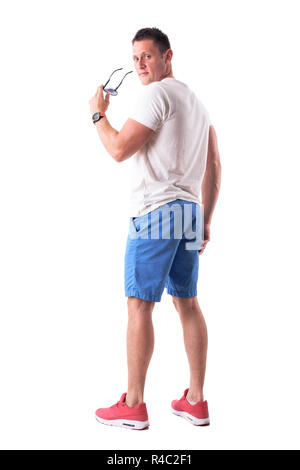 Modèle de mode confiant dans les vêtements d'été tenue des lunettes de soleil et à la recherche Retour à l'appareil photo. Vue arrière. Corps plein isolé sur fond blanc. Banque D'Images