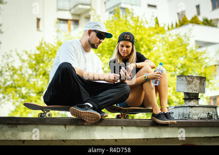 Garçon et fille sur une planche à roulettes sont assis et regarder quelque chose sur le téléphone mobile un jour d'été Banque D'Images