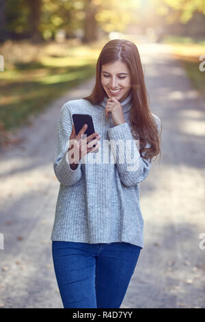 Jeune femme brune en chandail gris et bleu jeans en utilisant big smartphone dans boîtier noir, tout en descendant la route de campagne. En regardant l'écran et smi Banque D'Images