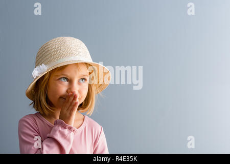 Adorable petite fille de quatre ans portant un élégant chapeau de paille whispering a secret avec sa main à sa bouche sur blue with copy space Banque D'Images