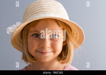 Adorable petite fille blonde de quatre ans avec d'énormes yeux verts souriant joyeusement à l'appareil photo portant un chapeau de paille à la mode Banque D'Images