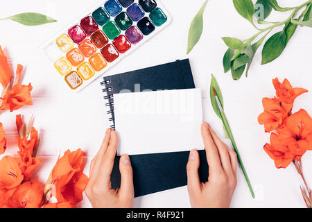 Woman's hands holding petite carte vierge près de peintures aquarelle, pinceau, fleurs et feuilles rouges sur fond blanc, vue du dessus. Banque D'Images