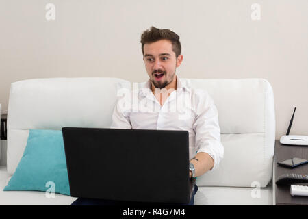 Homme avec portable. Handsome businessman working with laptop in office. Heureux jeune homme en chemise blanche assise sur un canapé à la maison, travailler sur ordinateur portable, en souriant. Banque D'Images