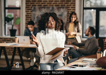 L'accent african american casual businesswoman holding journal avec des collègues qui travaillent derrière en bureau loft Banque D'Images