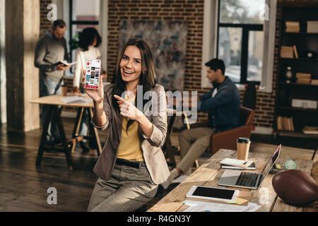 Smiling casual businesswoman holding smartphone avec l'application de pinterest dans loft bureau avec des collègues qui travaillent derrière Banque D'Images