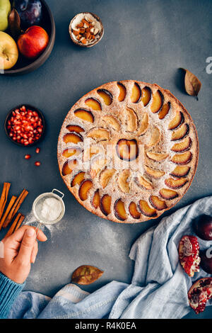 Une main le tamisage du sucre en poudre sur une pomme gâteau aux prunes photographié sur un fond foncé en vue de dessus. Banque D'Images