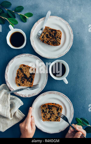 Mains prêt à manger une tranche de pain aux bananes et aux grains de chocolat sur une assiette blanche. Deux autres tranches sur deux assiettes et verres à accompagner. Banque D'Images