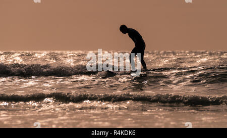 Surfer fatigué debout sur une planche de surf en provenance de la plage, à la fin d'une journée chaude, avec le soleil bas sur l'eau pétillante. Banque D'Images