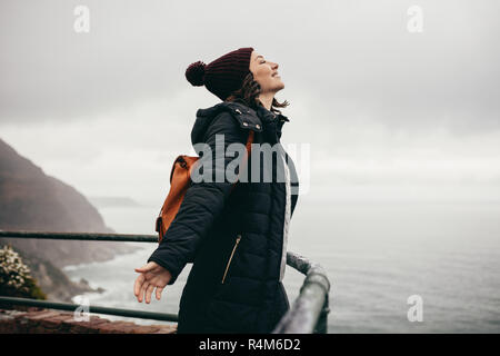 Femme portant veste chaude et hat standing by railing en montagne avec ses yeux fermés et les bras tendus. Woman standing in les entreprises fres Banque D'Images