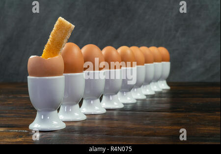 Rangée d'œufs durs dans white egg cups sur une table en bois le premier oeuf est ouverte avec un toast soldat plongé dans c Banque D'Images