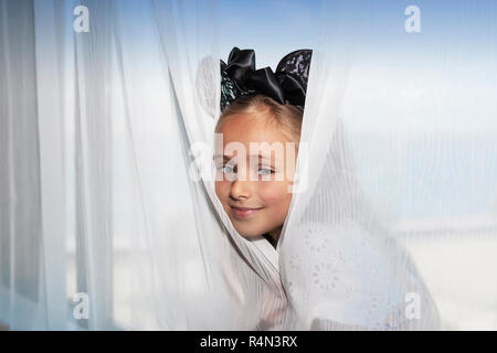 La petite fille est assise sur la fenêtre et à la recherche de derrière les rideaux. Banque D'Images