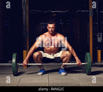 Bel homme assis brutal concentré et la préparation à la levée en Haltères fitness crossfit sur arrière-plan foncé sport club avec les ombres. Section de basse Banque D'Images