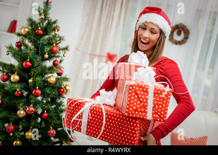 Belle jeune femme gaie wearing santa hat holding et de nombreux cadeaux de Noël à la maison. Banque D'Images