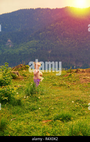 Cute young little girl blowing dandelion dans le coucher du soleil la lumière. Belle petite fille en rose chemise avec queue de peluche holding taraxacum de soufflage Banque D'Images